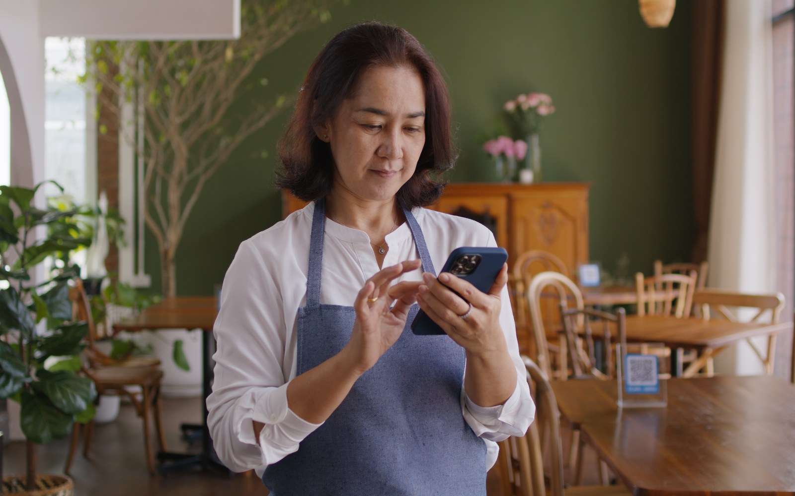 A restaurant owner in an apron checking bookings on her phone inside her restaurant
