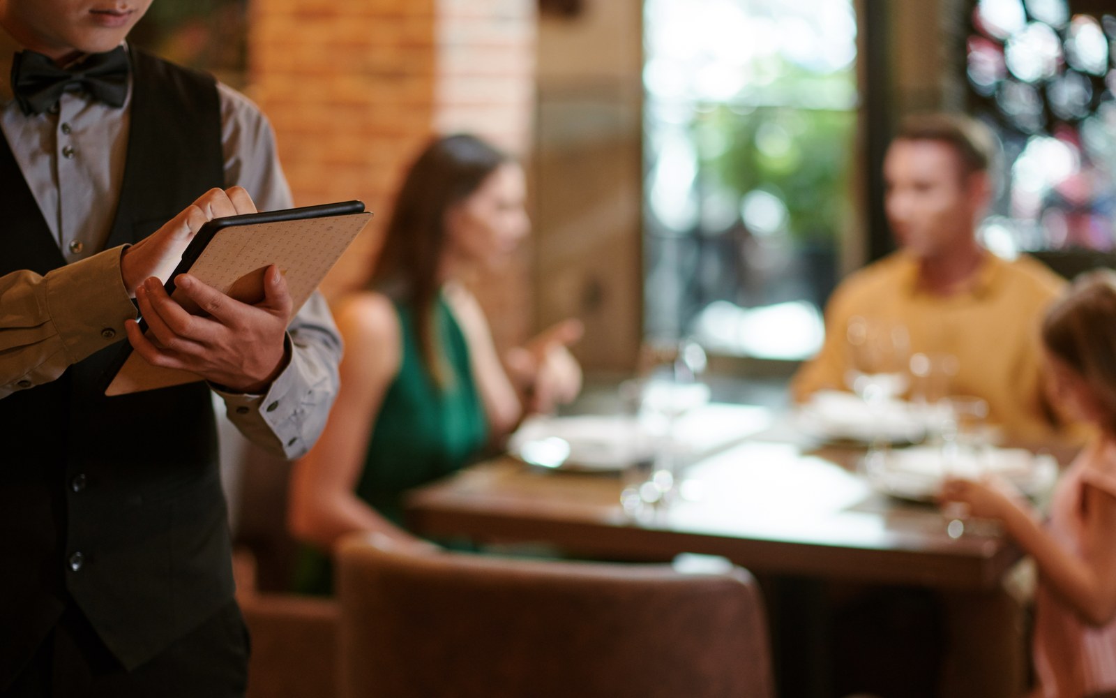 A waiter taking a booking on a handheld device from guests seated at a table in a warm restaurant